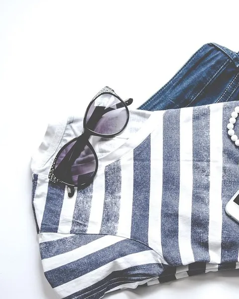 A flat lay of a striped shirt, sunglasses and a phone on a pale surface.